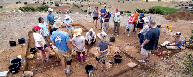 People at an archaeological dig stand and listen to someone squatting down in the work area